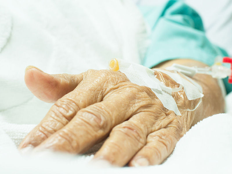 Mano geriátrica arrugada sobre una colcha blanca. Lugar de punción en el dorso de la mano. La línea conectada con pinza deslizante blanca se fija con esparadrapo blanco. El brazo lleva ropa hospitalaria verde desde el codo hacia abajo.