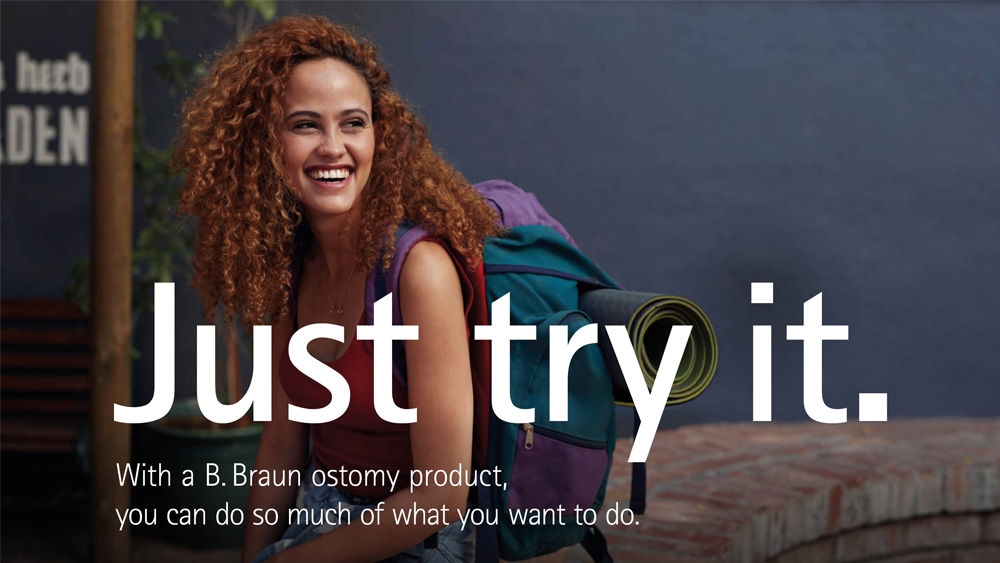 A woman with curly hair, wearing a red tank top and denim shorts, sits on a brick bench outdoors. She has a large backpack with a rolled-up yoga mat attached to it. There is a potted plant and part of a sign in the background. The text on the image reads Just try it. With a B. Braun ostomy product, you can do so much of what you want to.