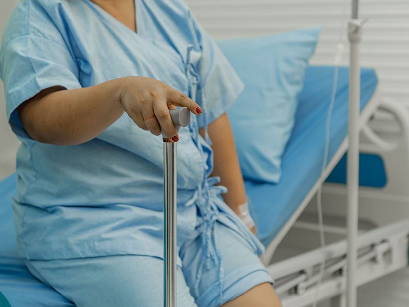 Paciente obesa de mediana edad con ropa azul de hospital sentada en el borde de una cama (cubrecama azul). La mano derecha sostiene un bastón para caminar. Hay un parche blanco en el antebrazo izquierdo. Hay una infusión junto a la cama.