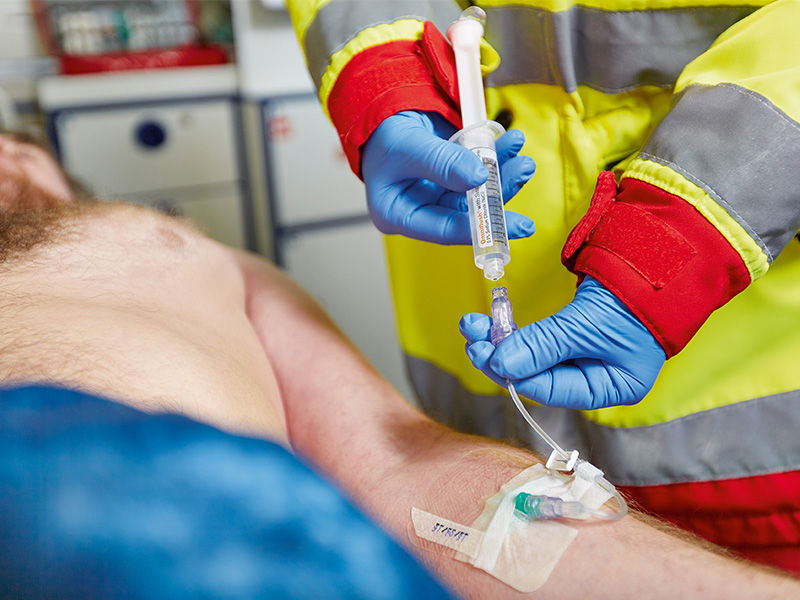 El paciente cubierto con una manta azul desde el centro del abdomen está tumbado dentro de una ambulancia. El antebrazo izquierdo del paciente se perfora con una cánula verde Introcan Safety® 3. La válvula Caresite con línea de infusión y pinza deslizante blanca está conectada. Fijación con esparadrapo para cánulas. Una persona con chaqueta de emergencia y guantes azules sostiene la jeringa llena en la mano derecha. La mano izquierda sostiene la línea debajo de la válvula.