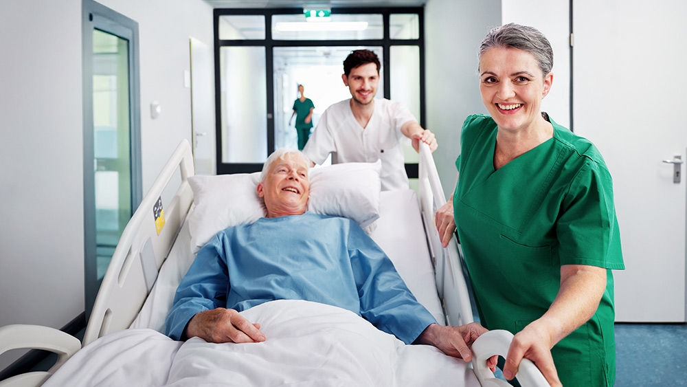 Paciente tumbado en una cama de hospital transportado por personal sanitario a través de un pasillo, con otro miembro del personal caminando a su lado