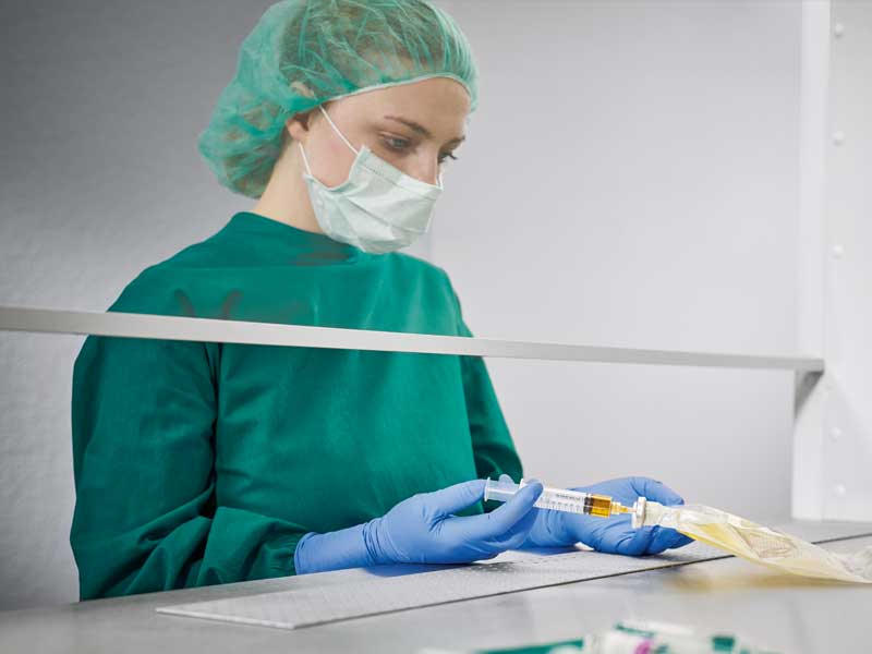 Una enfermera con uniforme estéril y mascarilla prepara la nutrición parenteral en un entorno estéril inyectando un líquido en una bolsa de nutrición. En primer plano se ven varios suministros médicos.