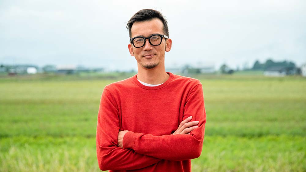 Una persona con gafas se encuentra de pie con confianza y brazos cruzados en un campo vasto. 